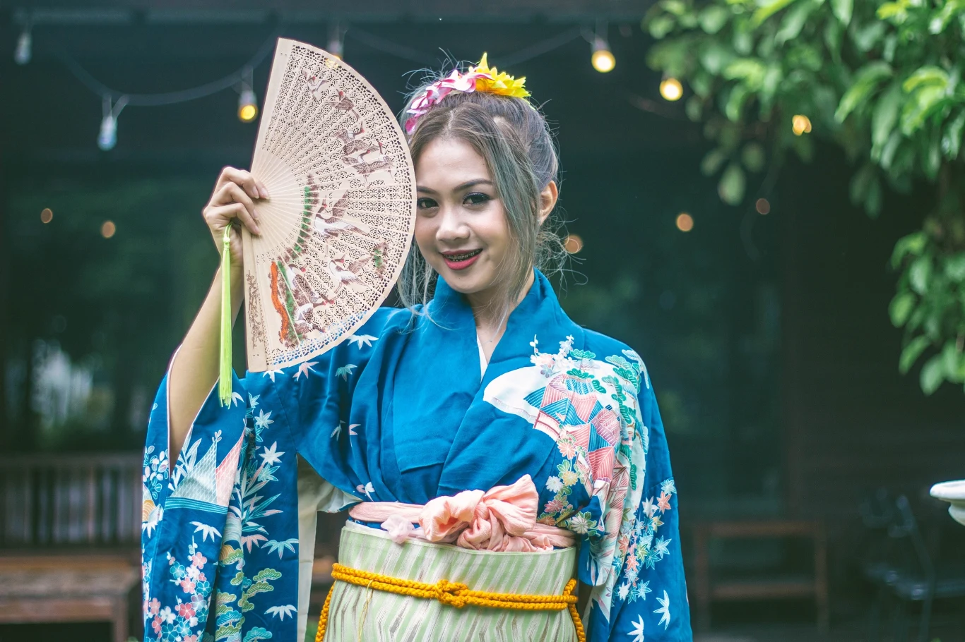 Kiedyś Japonki chłodziły się wachlarzami, dziś zastąpiły je podręczne wiatraczki. Młoda kobieta w tradycyjnym japońskim kimonie z kolorowym wzorem, trzymająca ozdobny wachlarz, z uśmiechem na twarzy i kwiatową ozdobą we włosach.