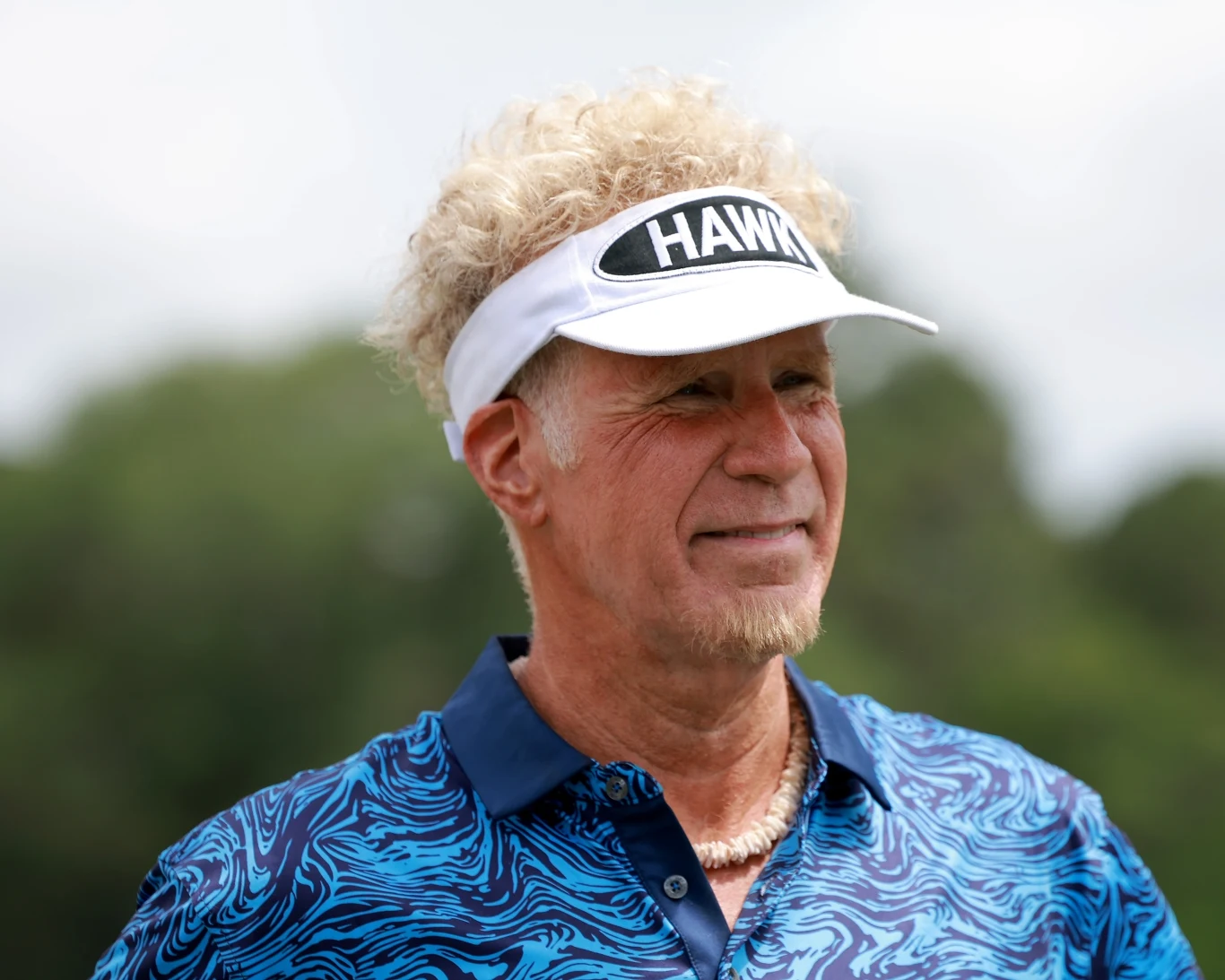 Will Ferrell na planie seriali "The Hawk" Mężczyzna z jasnymi, kręconymi włosami, ubrany w niebieską koszulkę z wzorem i biały sportowy daszek z napisem „HAWK”, na szyi ma naszyjnik z muszelek.