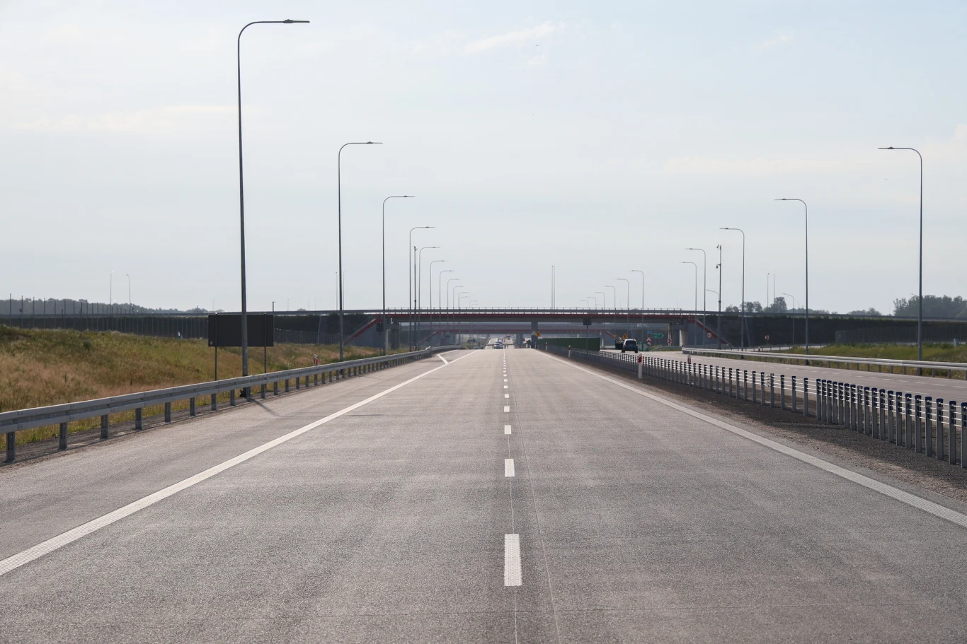 Szeroka, nowa jezdnia autostrady prowadząca w stronę wiaduktu, otoczona metalowymi barierkami i równomiernie rozmieszczonymi latarniami po obu stronach drogi.