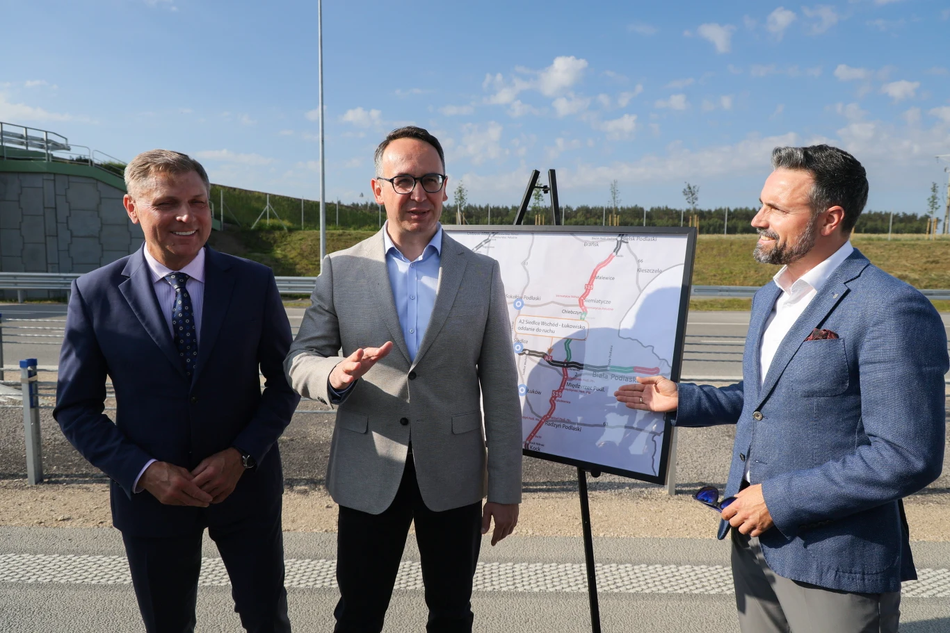 Trzech elegancko ubranych mężczyzn stoi na drodze obok planszy z mapą trasy komunikacyjnej, jeden z nich wskazuje na mapę, pozostali uśmiechają się do kamery.