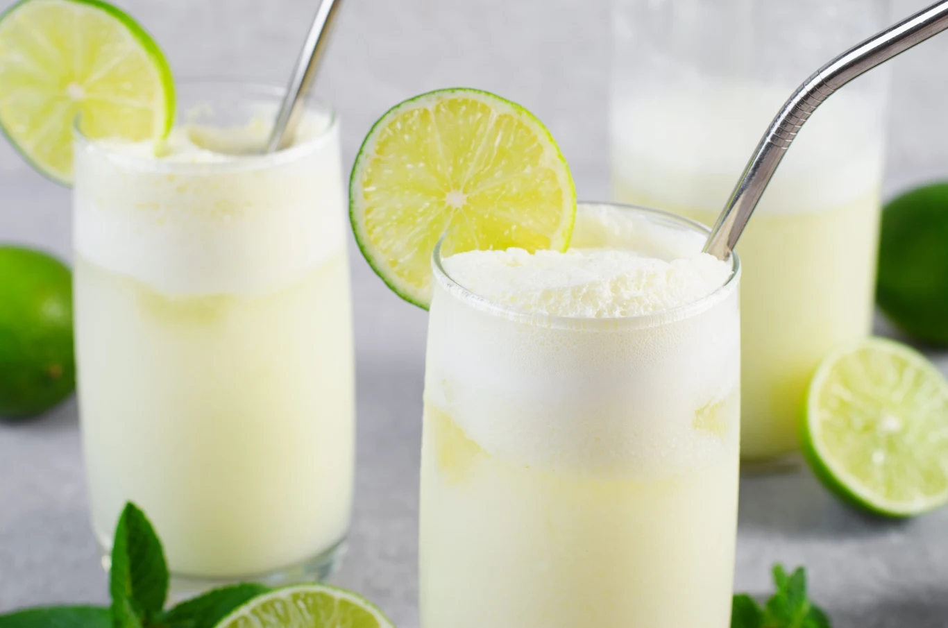 Szklanki wypełnione kremowym, jasnożółtym napojem udekorowanym plasterkami limonki i metalowymi słomkami, otoczone świeżymi limonkami i listkami mięty.