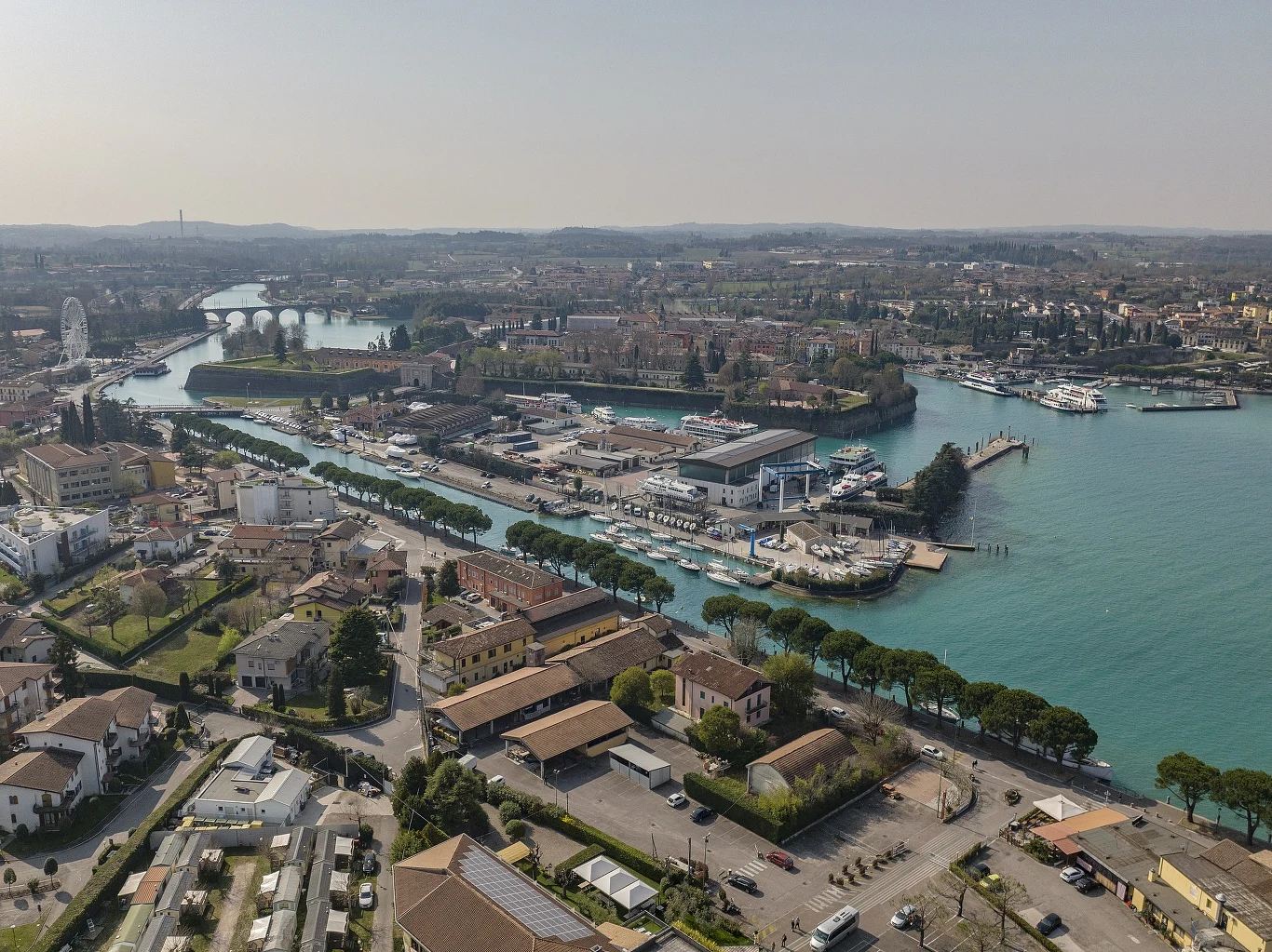 Rozległy port z mariną i cumującymi jachtami, otoczony niską zabudową mieszkalną oraz szerokimi alejami drzew prowadzącymi wzdłuż brzegu wody.