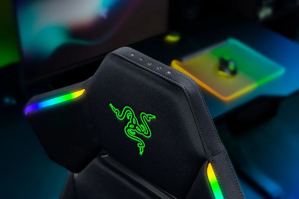 Gamingowy fotel z podświetlanymi, wielokolorowymi elementami LED oraz wyraźnym neonowym logo marki Razer na oparciu, ustawiony przy nowoczesnym stanowisku komputerowym.