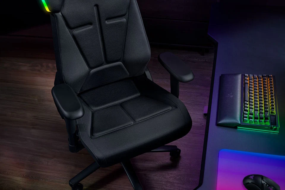 Czarny, ergonomiczny fotel gamingowy z podłokietnikami, ustawiony obok biurka z mechaniczną klawiaturą podświetlaną wielobarwnym światłem.