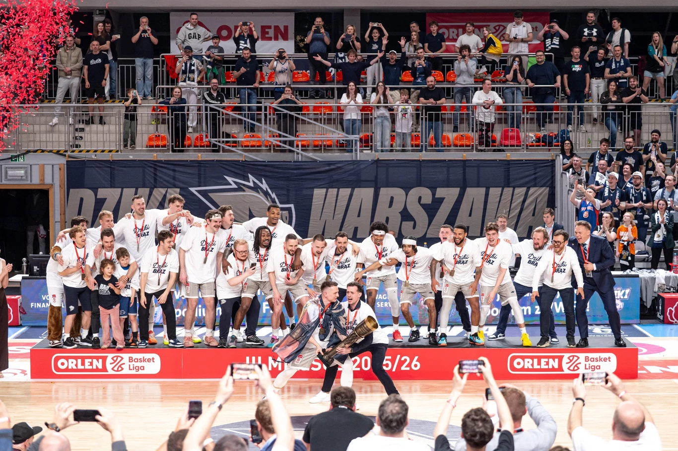 Dziki Warszawa. Jak w mieście Legii zbudowano fenomen od zera Koszykarze drużyny Legia Warszawa ubrani w białe koszulki świętują zdobycie mistrzostwa, otoczeni medalami i pucharem, zebrani na środku parkietu wśród wiwatujących kibiców i fotoreporterów.