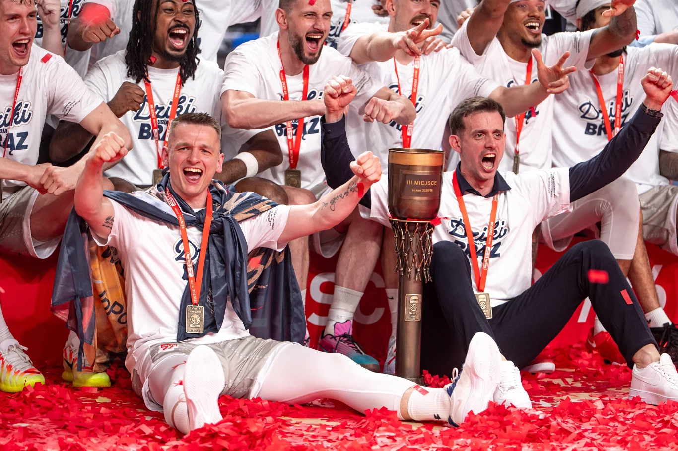 Dziki Warszawa wywalczyły historyczny medal Grupa uśmiechniętych zawodników koszykarskich w białych koszulkach świętuje zwycięstwo, siedząc wokół dużego pucharu, wśród konfetti i z medalami na szyjach.