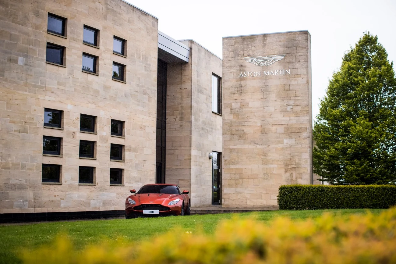 Czerwony samochód Aston Martin zaparkowany przed budynkiem z logo marki, otoczony zielonym trawnikiem i drzewami.