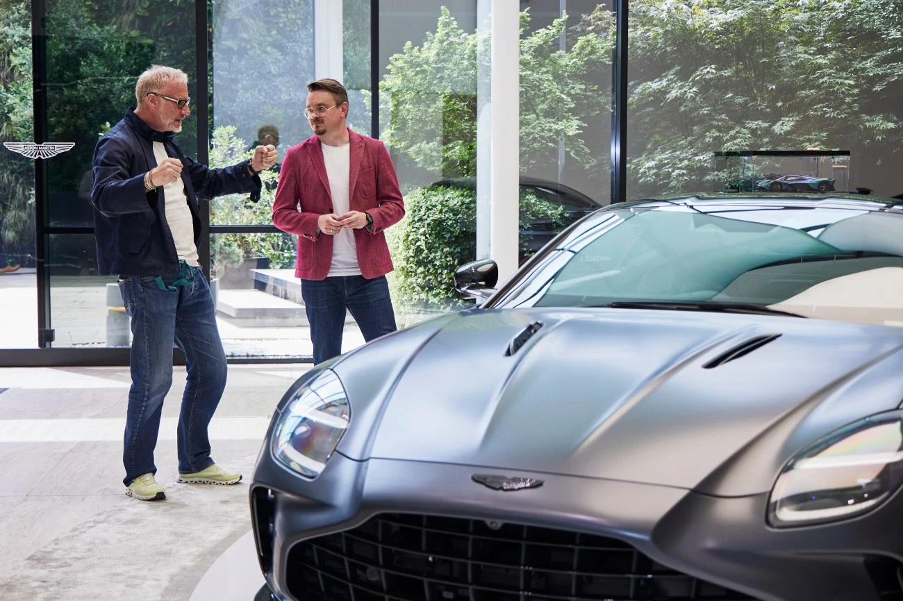 Dwóch mężczyzn rozmawiających obok luksusowego samochodu Aston Martin w nowoczesnym, jasnym salonie z dużymi oknami i widokiem na zieleń za szybą.