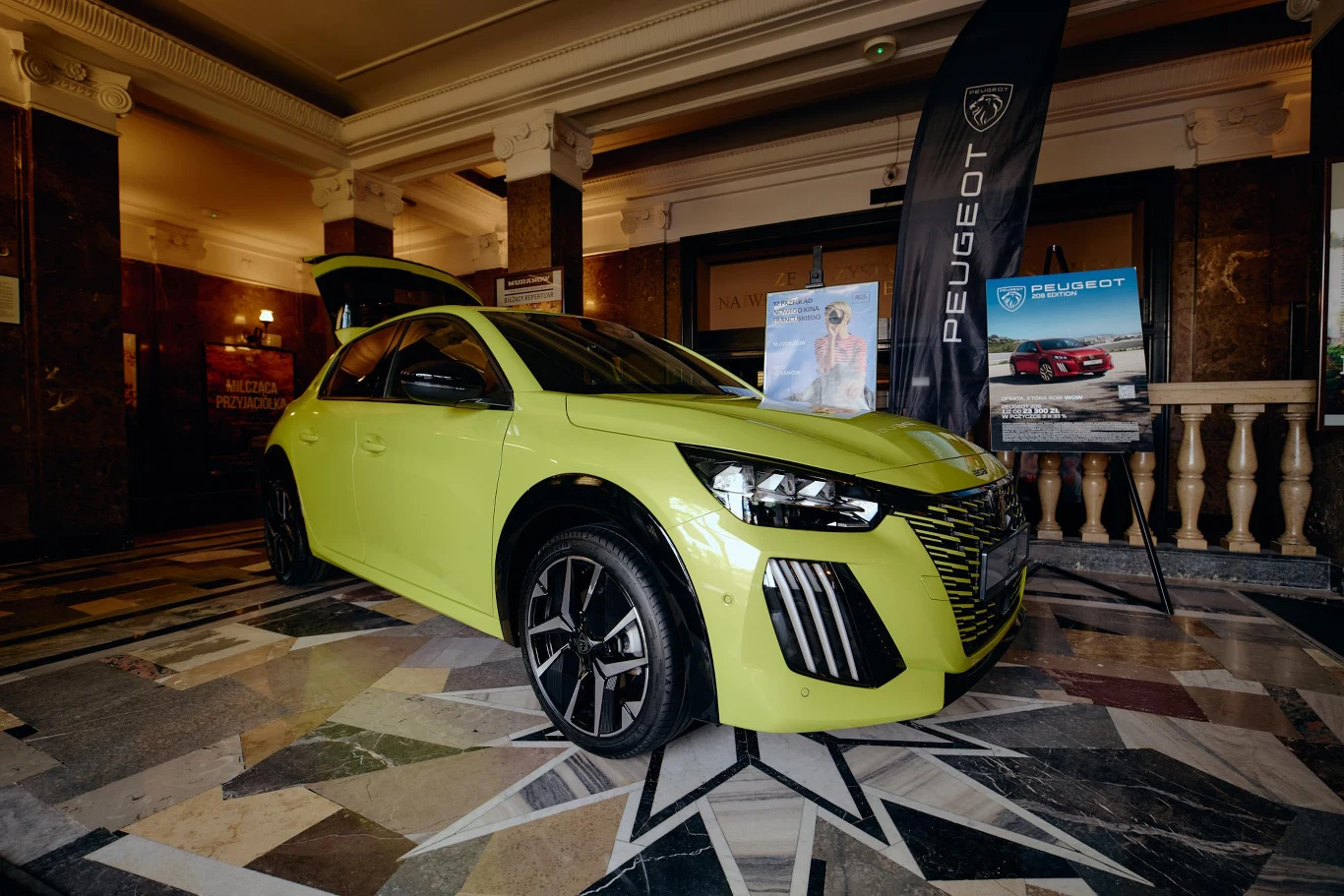 Żółtozielony samochód Peugeot ustawiony na marmurowej posadzce wnętrza budynku, obok stoją dwa roll-upy z logo i grafikami marki.
