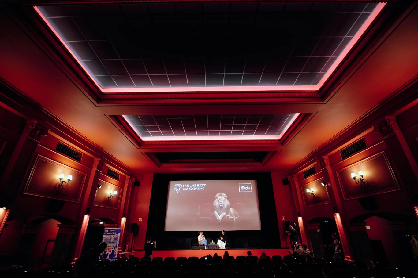 Kino z czerwonymi ścianami, dużym ekranem wyświetlającym zbliżenie osoby z mikrofonem i niewielką grupą osób na scenie przed publicznością.