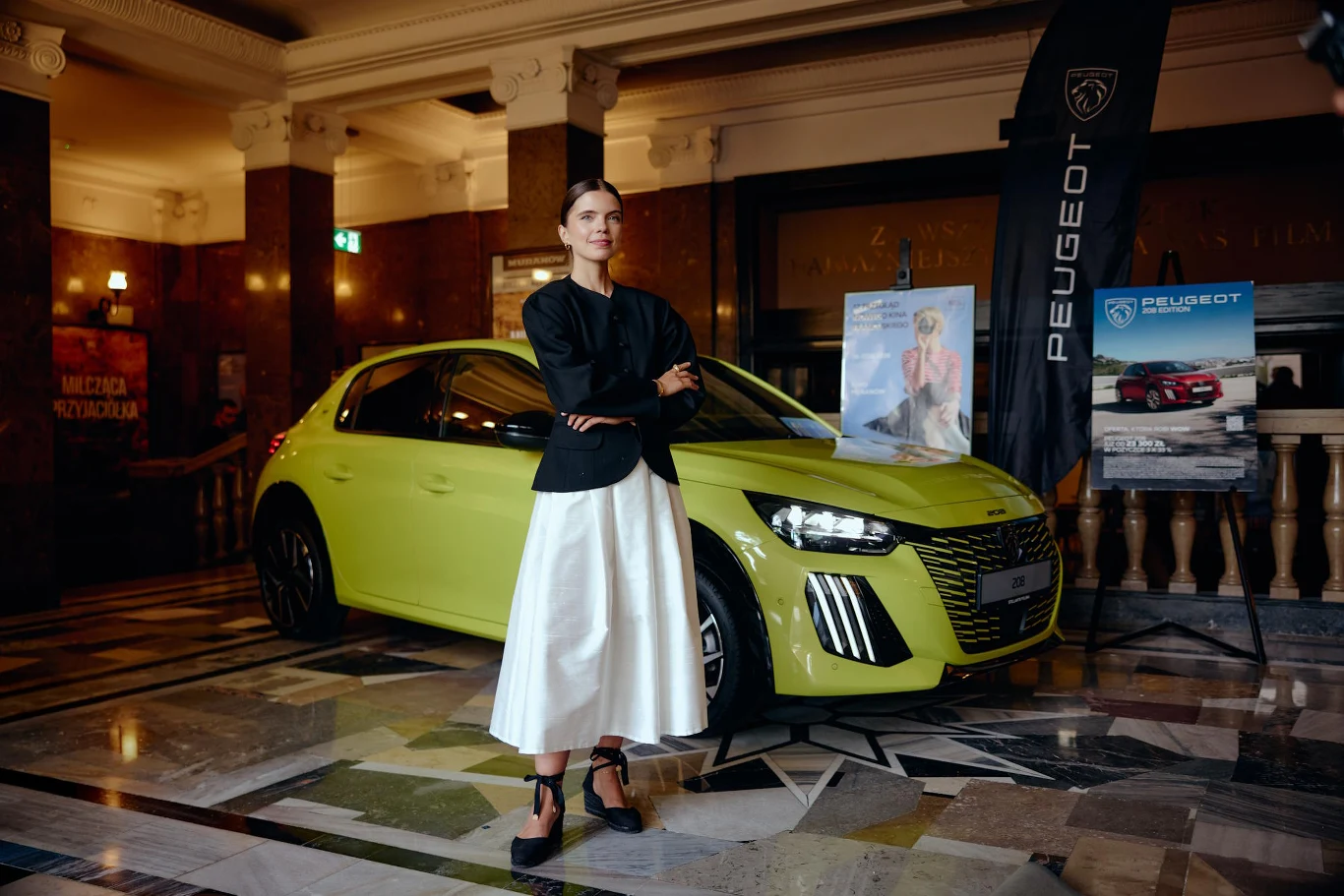 Stylowo ubrana kobieta stojąca przed żółtym samochodem Peugeot w eleganckim, marmurowym wnętrzu, obok stojaki z materiałami promocyjnymi marki.