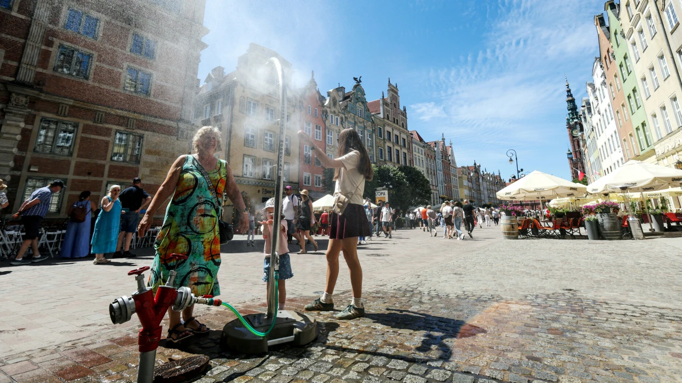 Dwie kobiety chłodzą się pod miejską kurtyną wodną rozstawioną na brukowanym placu w otoczeniu zabytkowych kamienic i spacerujących przechodniów.