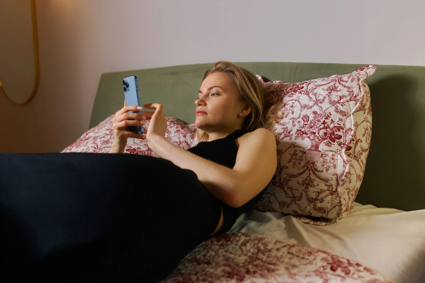 Kobieta w czarnym topie leży na łóżku z poduszkami w czerwone wzory, trzymając telefon i patrząc w ekran.