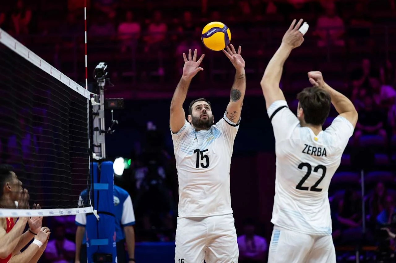 Luciano De Cecco, rozgrywający reprezentacji Argentyny Dwóch siatkarzy w białych strojach, jeden z numerem 15 wykonuje wystawienie piłki, a drugi z numerem 22 przygotowuje się do ataku przy siatce podczas meczu halowego.