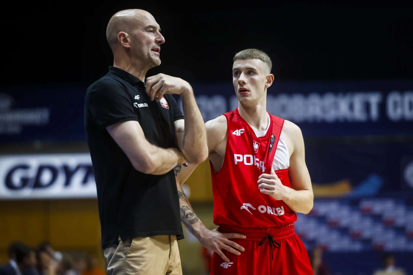 Jakub Andrzejewski Trener w czarnej koszulce i zawodnik w czerwonym stroju koszykarskim z napisem 'Polska' intensywnie rozmawiają podczas przerwy w meczu.