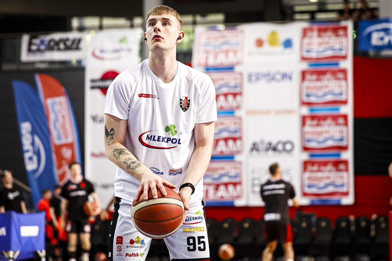 Jakub Andrzejewski Koszykarz w białym stroju z piłką przygotowujący się do rzutu na hali sportowej, w tle rozmyci zawodnicy i bannery sponsorów.