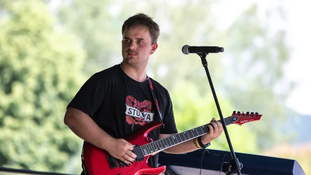 Mężczyzna grający na czerwonej gitarze elektrycznej, stojący przy mikrofonie na scenie obok klawiatury.