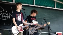 Dwóch młodych muzyków grających na gitarach elektrycznych i śpiewających na scenie podczas koncertu na świeżym powietrzu, w tle perkusja oraz sprzęt muzyczny.