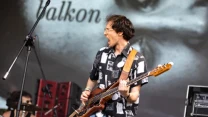 Mężczyzna w koszuli z krótkim rękawem gra na gitarze basowej na scenie, obok widoczny fragment zestawu perkusyjnego i sprzętu nagłośnieniowego, w tle ekran z napisem.