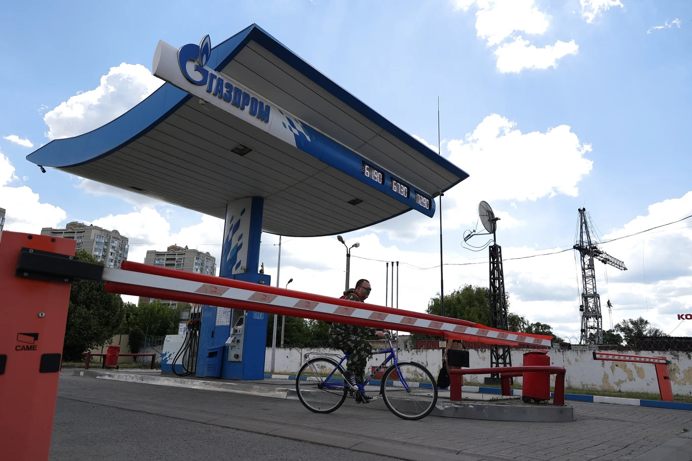 Wiele stacji zostało całkowicie zamkniętych, bo nie mają co sprzedawać Rowerzysta przeprowadza rower przez szlaban na tle nieczynnej stacji benzynowej Gazpromu, a w oddali widoczny jest żuraw budowlany.