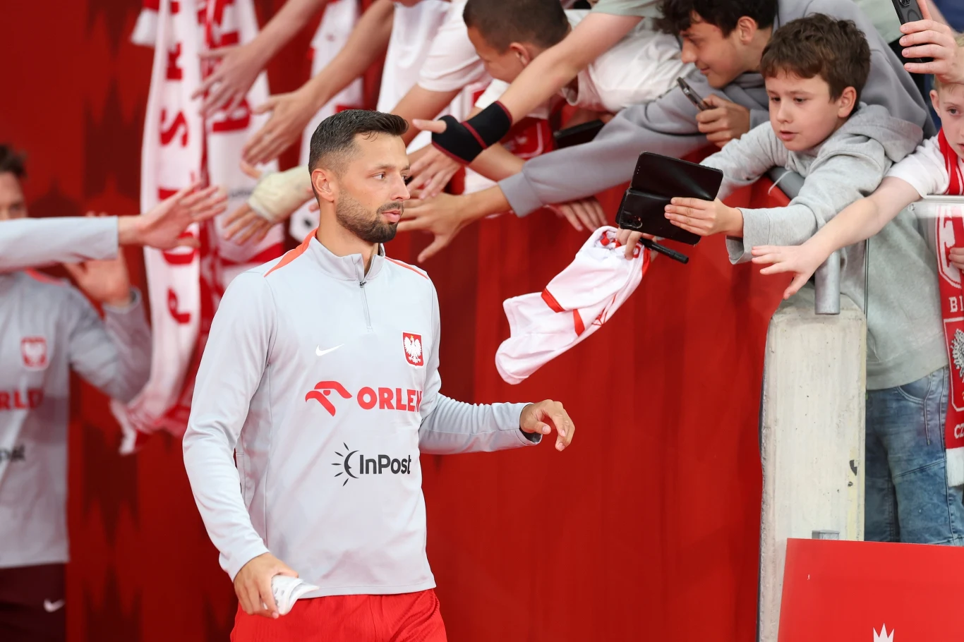 Piłkarz reprezentacji Polski wychodzi ze strefy tunelu, a kibice podają mu przedmioty do podpisu i wyciągają ręce, licząc na autograf.
