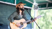 Brodaty mężczyzna w kapeluszu kowbojskim gra na gitarze akustycznej i śpiewa do mikrofonu na scenie plenerowej.