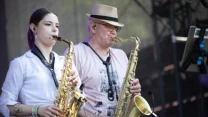 Dwójka muzyków grających na saksofonach, kobieta i mężczyzna stoją obok siebie na scenie koncertowej, widoczne mikrofony i stojak z nutami.
