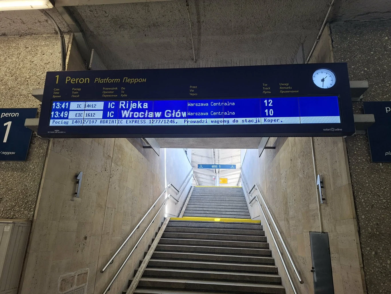 Elektroniczna tablica odjazdów z informacjami o pociągach Intercity do Rijeki oraz Wrocławia Głównego, umieszczona nad schodami prowadzącymi na peron, ściany z betonu i minimalistyczne oświetlenie.