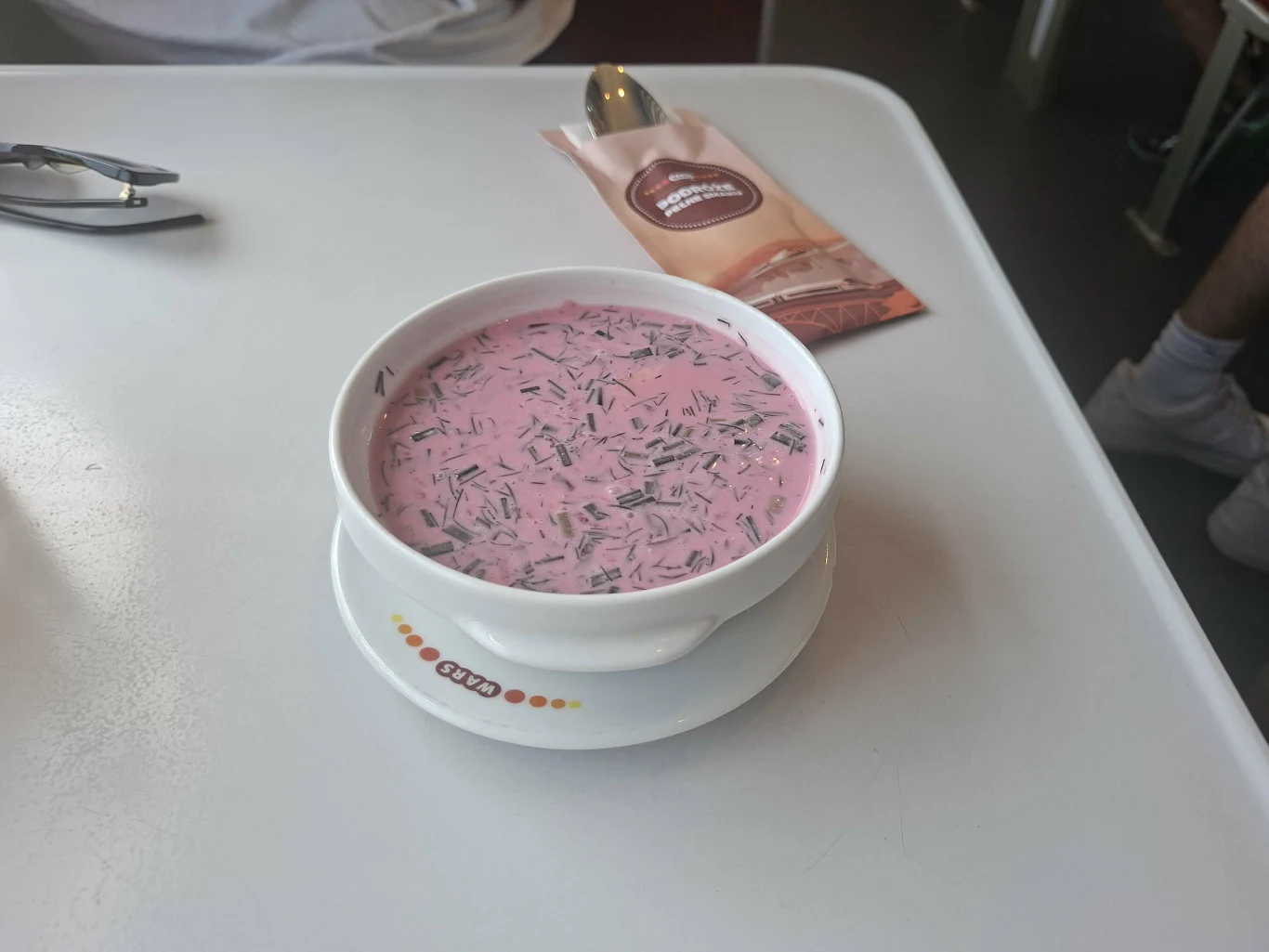 Biała miska z różową, chłodnikową zupą z dodatkiem posiekanego koperku, ustawiona na talerzyku na jasnym stole obok opakowania z łyżką.