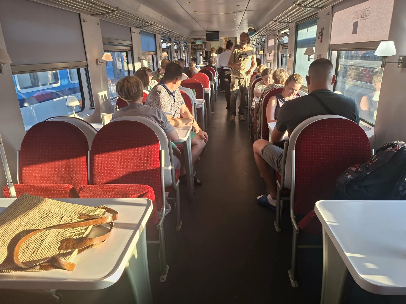 Wagon restauracyjny pociągu z czerwonymi, tapicerowanymi siedzeniami przy białych stolikach, pasażerowie siedzący i rozmawiający lub odpoczywający, na jednym ze stolików leży słomiana torba.