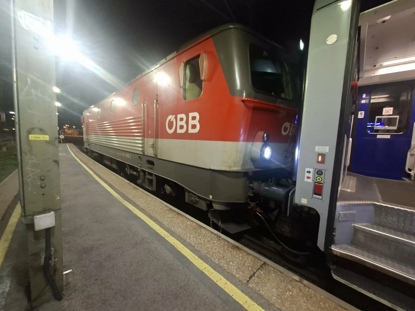 Czerwona lokomotywa kolejowa z oznaczeniem ÖBB stoi na peronie obok wejścia do nowoczesnego wagonu pasażerskiego oświetlonego światłem wewnętrznym.