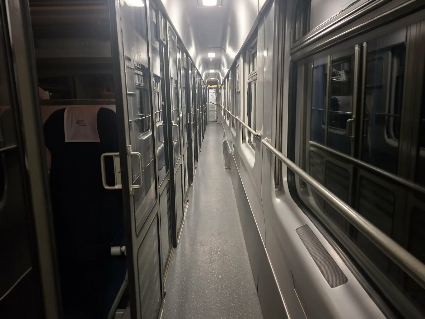 Wąski korytarz wagonu pociągu z zamkniętymi przedziałami po lewej i oknami po prawej stronie, widoczne jasne oświetlenie sufitowe.
