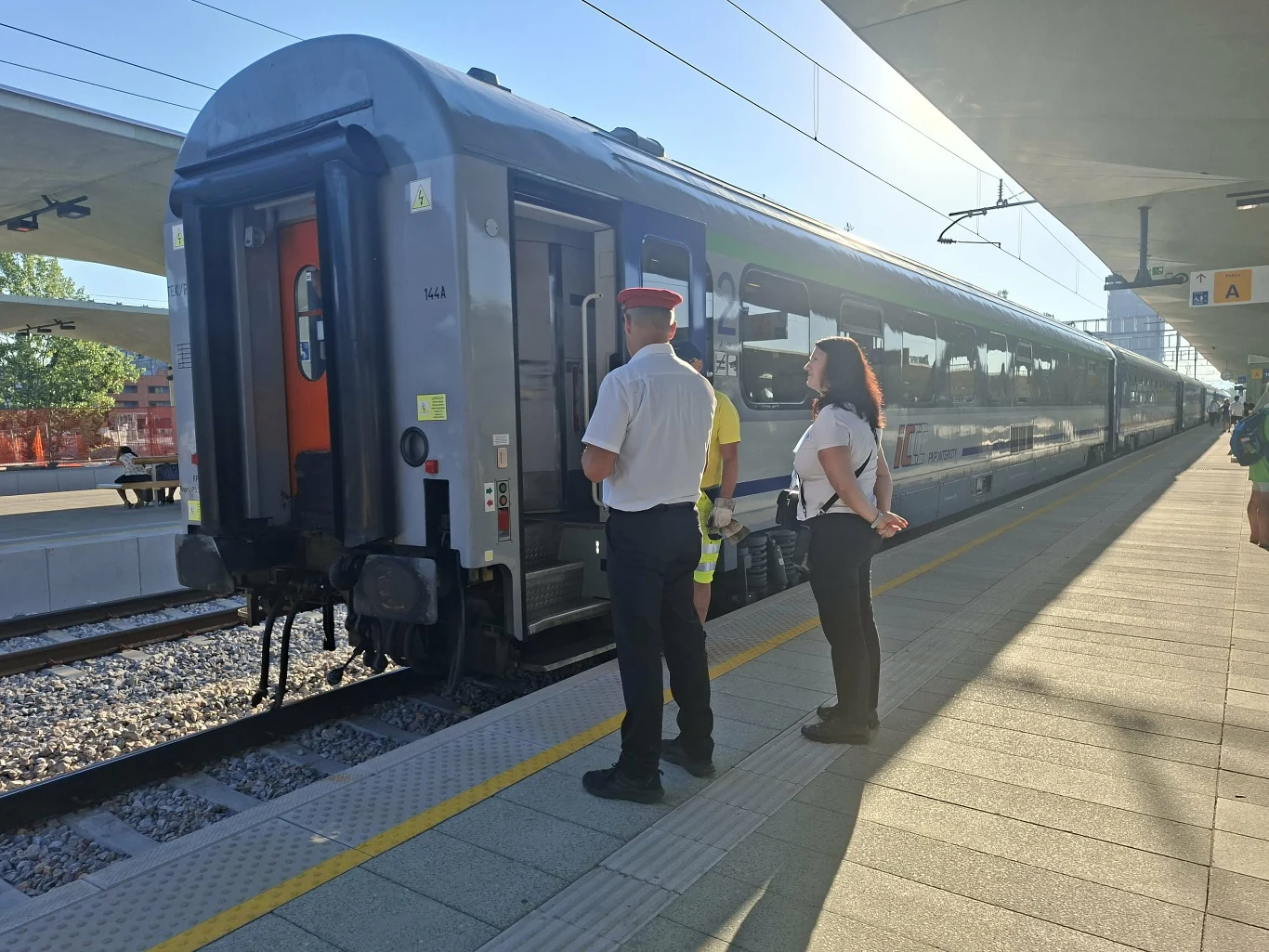 Ostatni wagon pociągu stojącego na peronie, obok którego znajdują się trzy osoby w służbowych uniformach kolejowych zwrócone w kierunku pociągu.