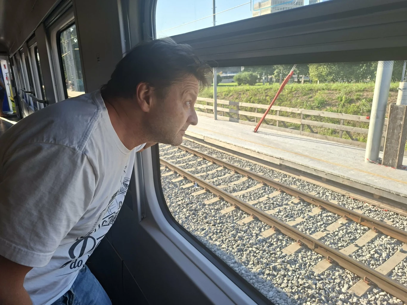 Mężczyzna w jasnej koszulce pochyla się i wygląda przez okno pociągu na mijane tory oraz platformę stacji kolejowej.