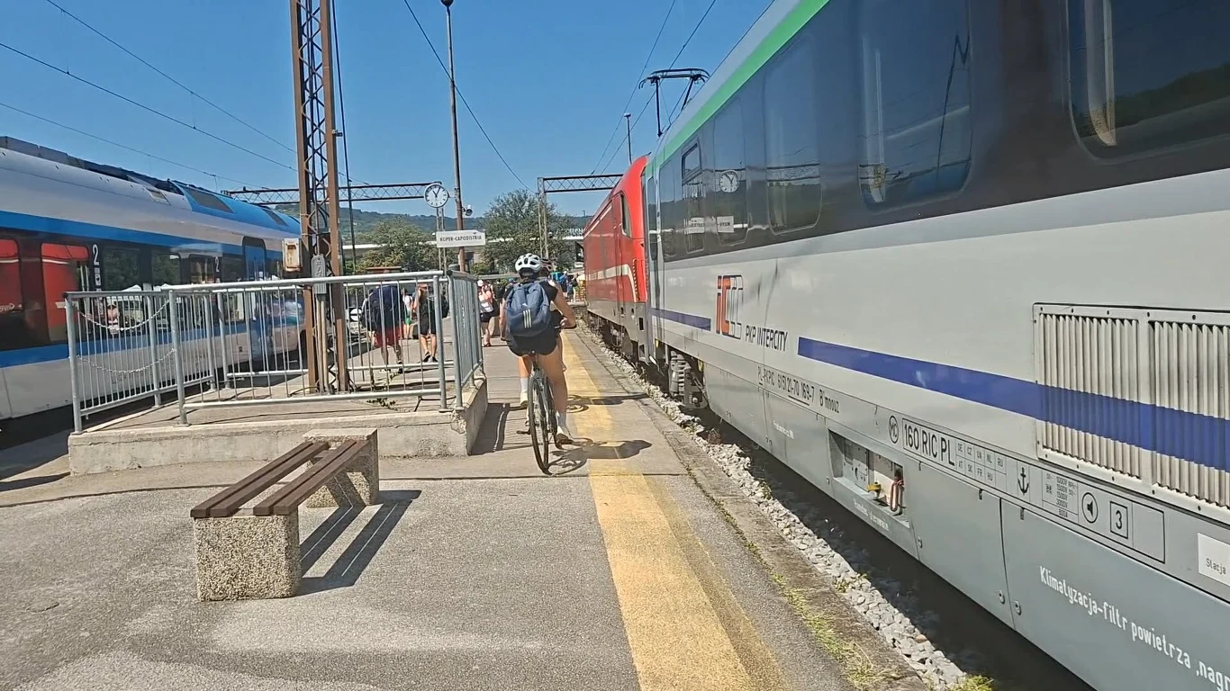 Rowerzysta w kasku przejeżdżający przez peron kolejowy obok dwóch stojących pociągów, w słoneczny dzień.