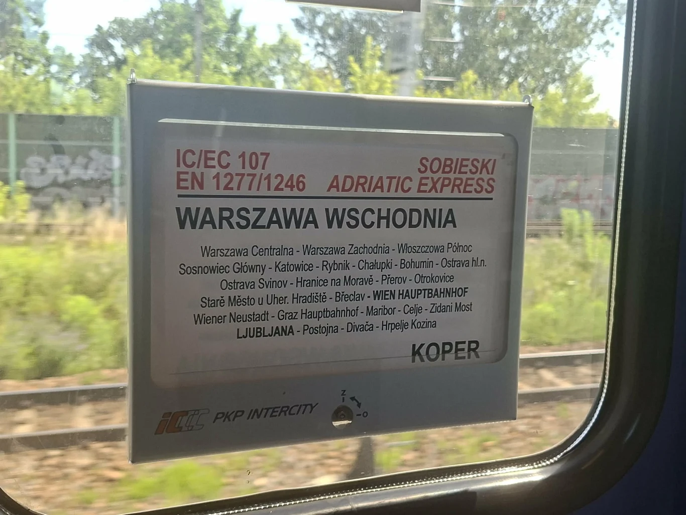 Tabliczka z rozkładem trasy pociągu IC/EC 107 Sobieski Adriatic Express relacji Warszawa Wschodnia – Koper, umieszczona na szybie pociągu, z widoczną listą stacji pośrednich wypisaną na białym tle.