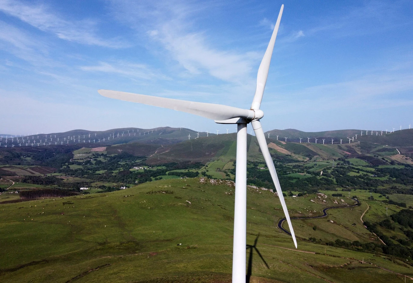 Duża turbina wiatrowa na tle zielonych wzgórz z rozproszonymi w oddali kolejnymi turbinami wiatrowymi, pod błękitnym niebem.