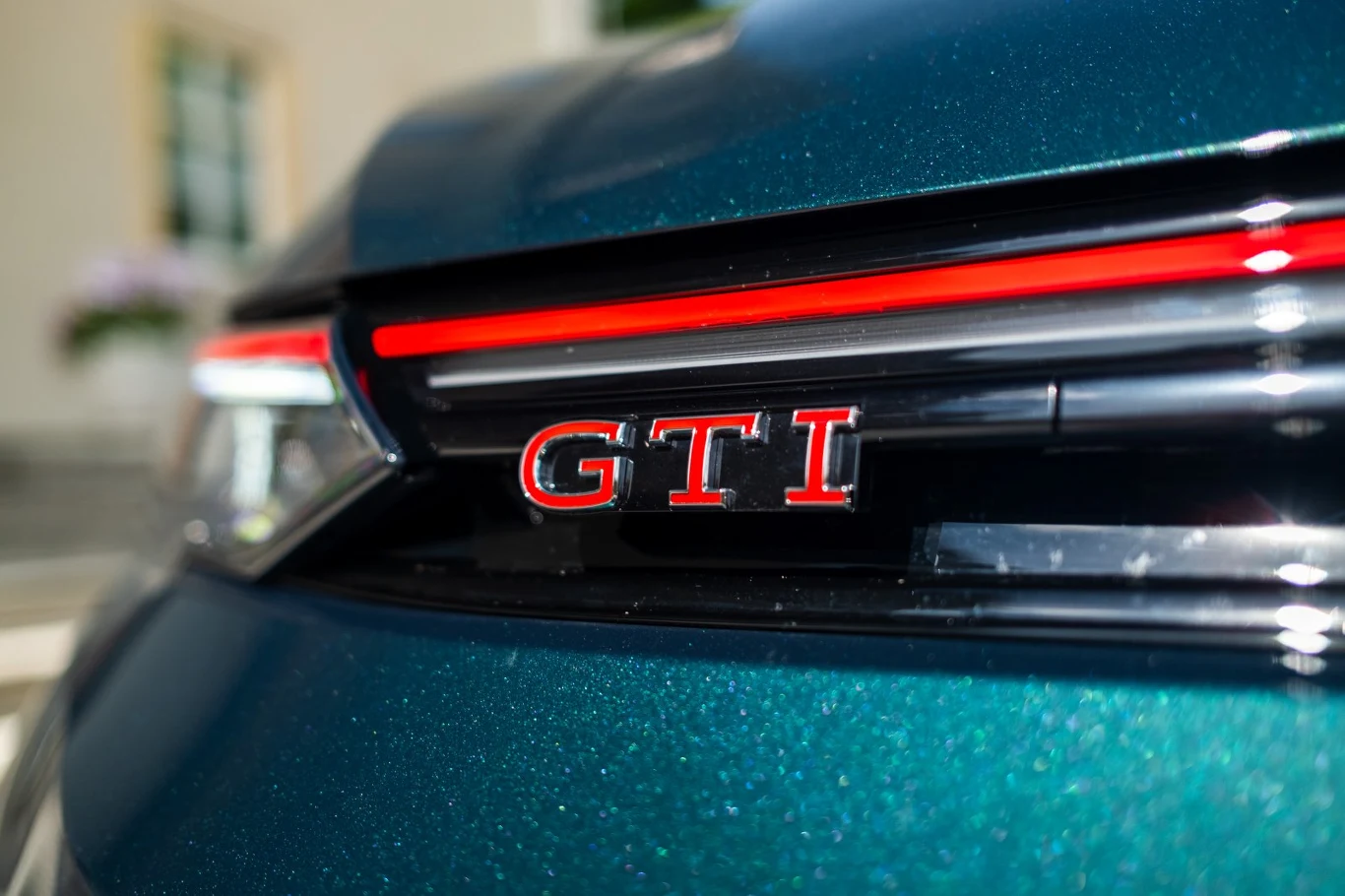 Czerwony emblemat GTI umieszczony na błyszczącej, ciemnozielonej karoserii samochodu, podkreślony poziomym czerwonym paskiem i nowoczesnymi liniami reflektora.