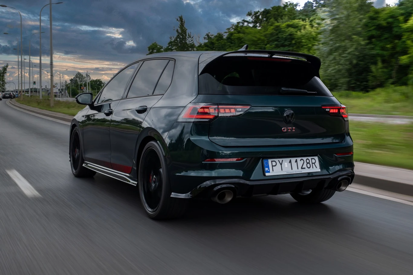 Czarny hatchback z oznaczeniem GTI na tylnej klapie jedzie lewym pasem drogi miejskiej, otoczony zielenią i podmiejską zabudową.