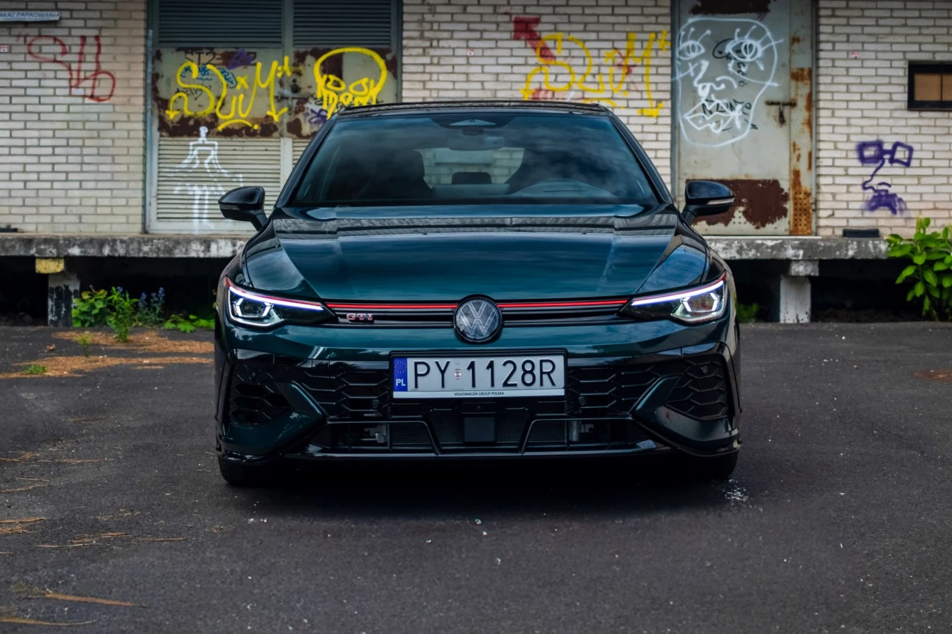 Ciemnozielony Volkswagen z charakterystycznym czerwonym akcentem na grillu, zaparkowany na tle zaniedbanego budynku z graffiti.