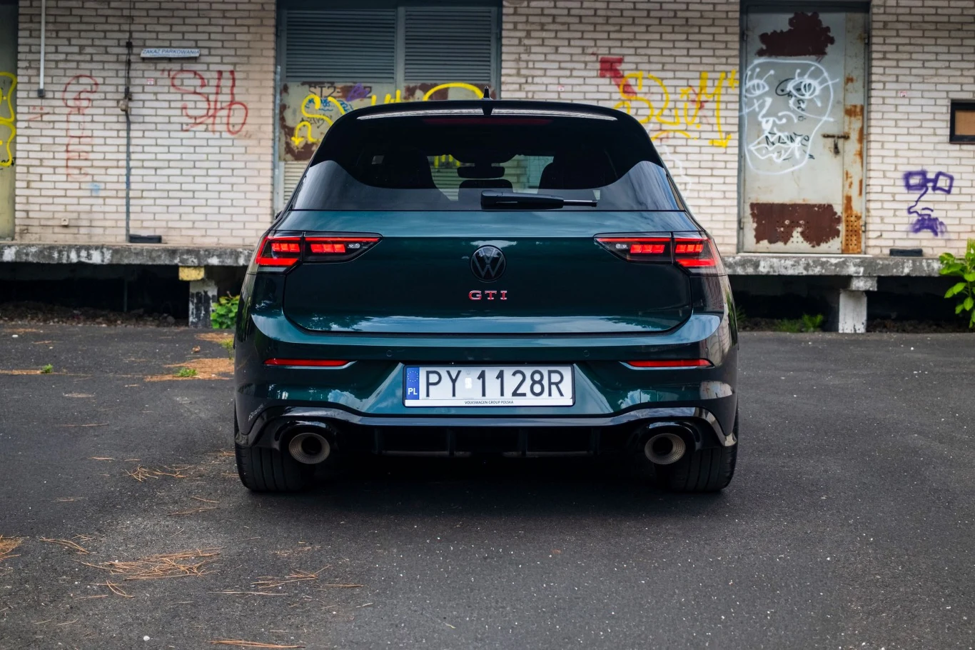 Ciemnozielony samochód hatchback z literami GTI na tylnej klapie, sportowa sylwetka, podwójny wydech i polska tablica rejestracyjna PY 1128R, w tle ceglana ściana z graffiti.