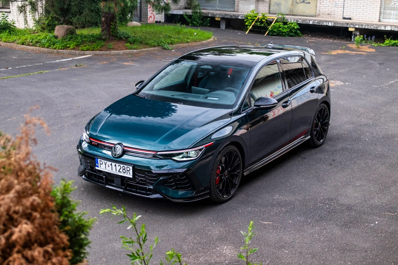 Ciemnozielony hatchback Volkswagen z czarnymi felgami i czerwonymi akcentami stoi na asfaltowej powierzchni, otoczony niską zielenią.