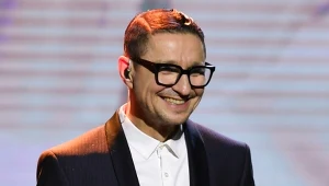 Uśmiechnięty mężczyzna w okularach i eleganckim garniturze z białą koszulą stoi na scenie, oświetlony reflektorami.