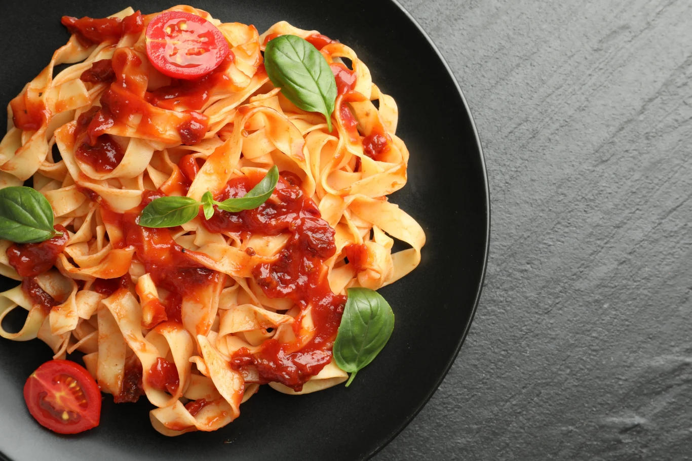 Makaron tagliatelle z sosem pomidorowym, kawałkami pomidorów i świeżymi listkami bazylii podany na czarnym talerzu.