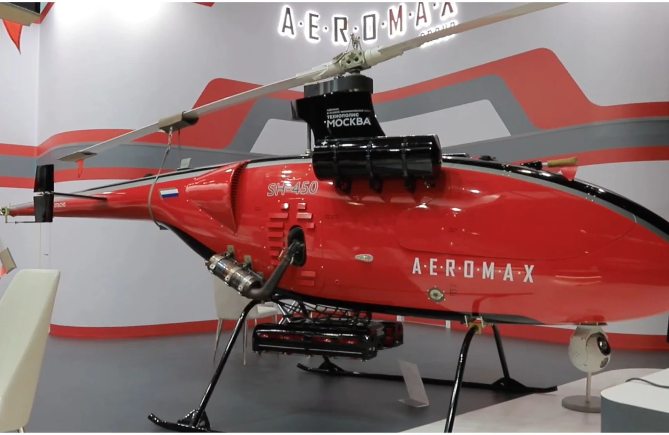 Czerwony bezzałogowy śmigłowiec z oznaczeniem AEROMAX i bocznym wirnikiem ustawiony na ekspozycji, wyposażony w mechaniczne podwozie oraz dodatkowy sprzęt zamontowany pod kadłubem.
