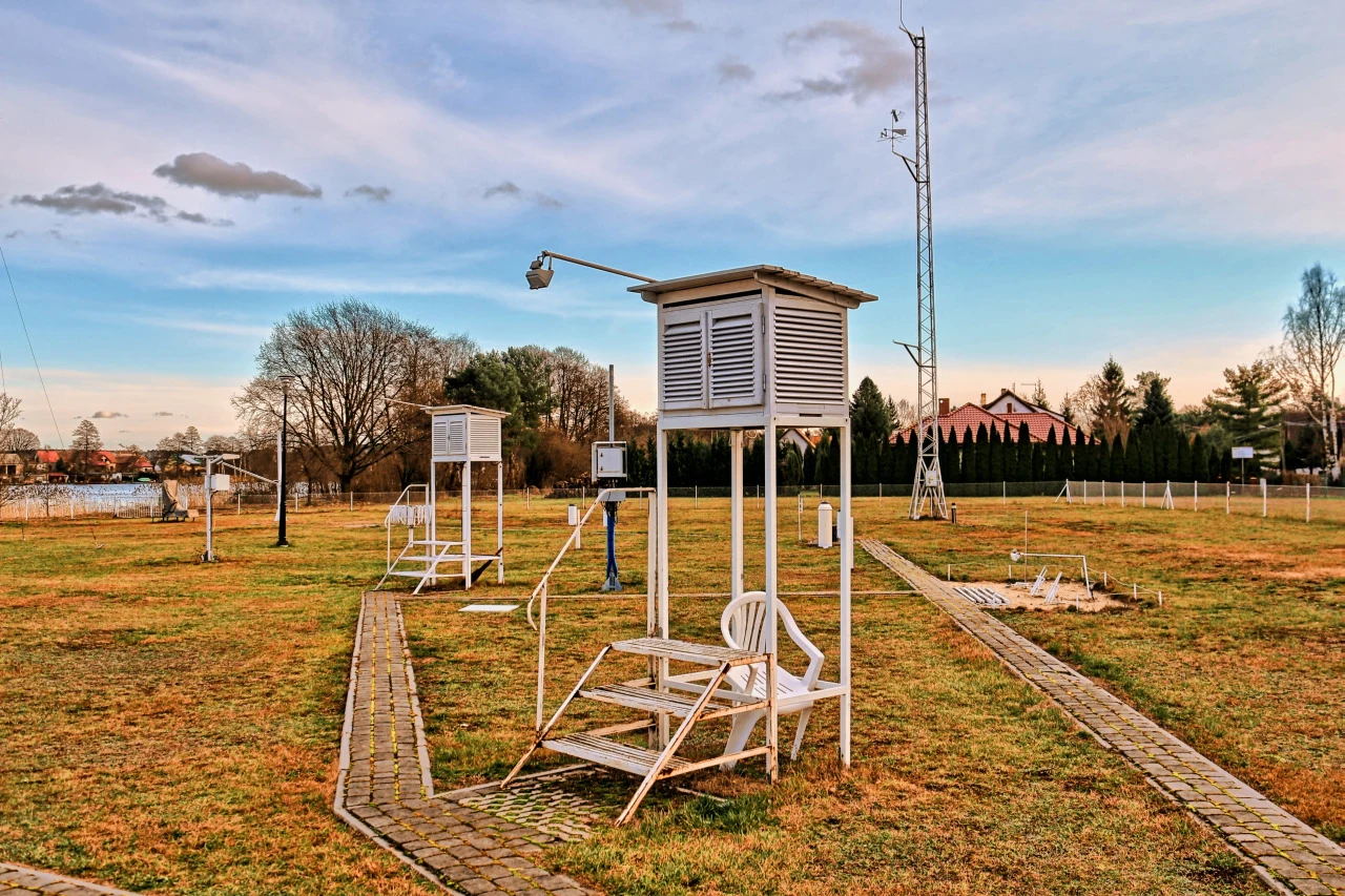 Stacja meteorologiczna z przyrządami pomiarowymi ustawionymi na trawiastej działce, z białą klatką meteorologiczną na pierwszym planie oraz wysokim masztem z czujnikami w tle.