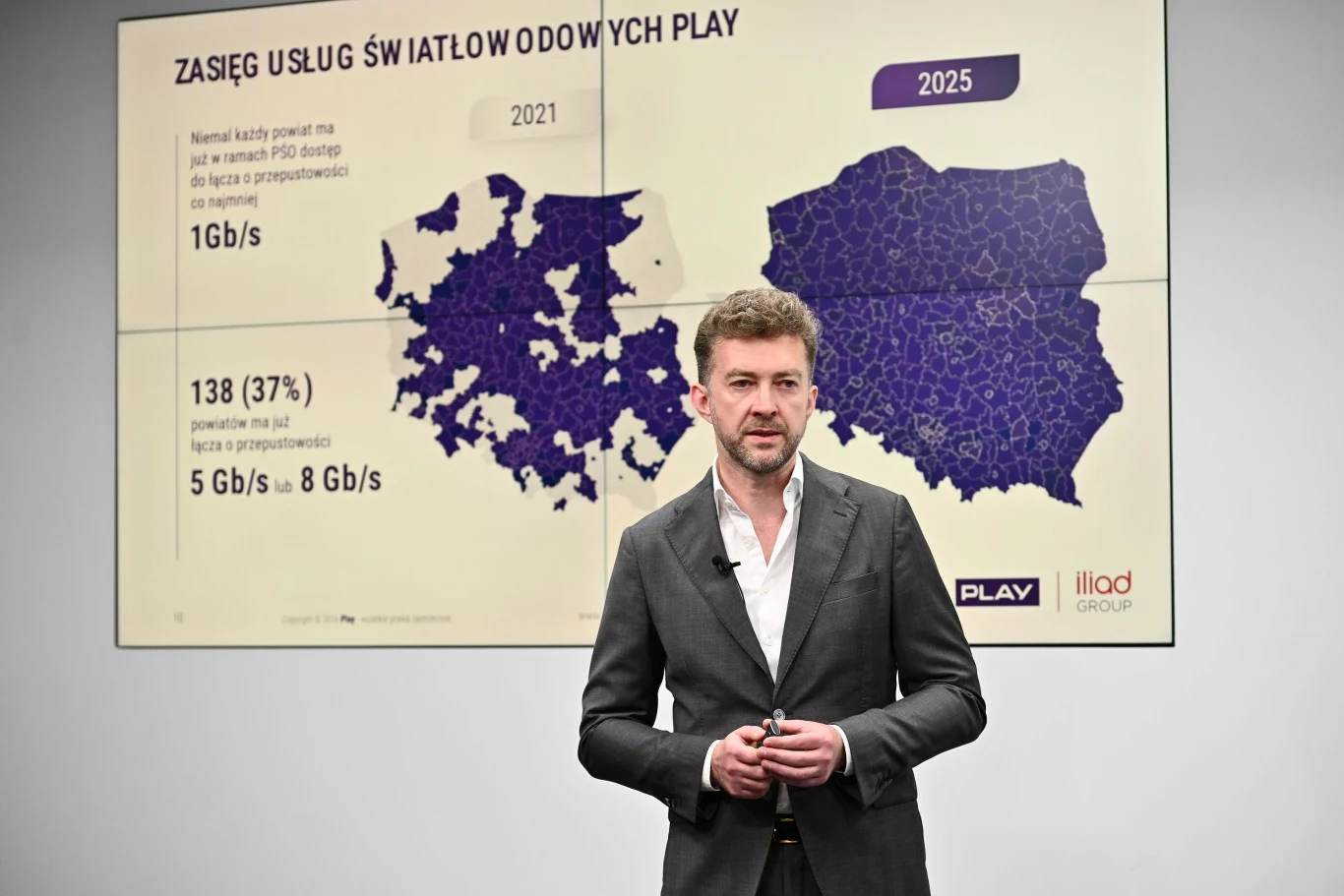 Mężczyzna w garniturze stoi przed tablicą z mapami Polski oraz danymi dotyczącymi zasięgu usług światłowodowych Play na lata 2021 i 2025.
