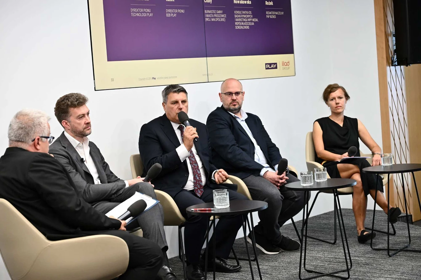 Panel dyskusyjny z udziałem pięciu osób siedzących w fotelach, jedna osoba mówi do mikrofonu, pozostali słuchają; na stolikach szklanki z wodą i notatniki, w tle fragment dużego ekranu z agendą.
