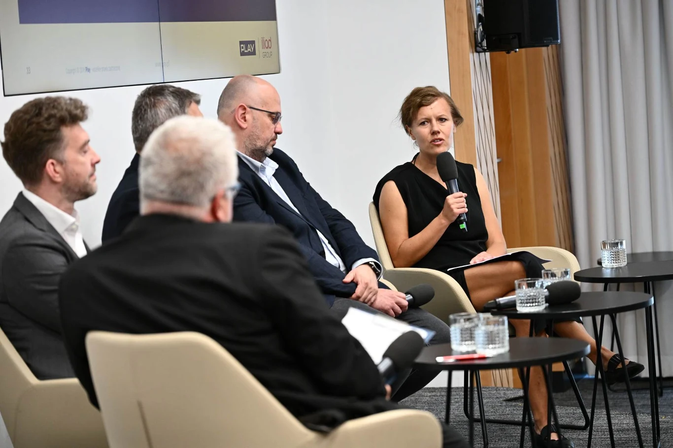 Panel dyskusyjny z udziałem czterech mężczyzn i jednej kobiety siedzących na krzesłach, kobieta trzyma mikrofon i przemawia, przed uczestnikami znajdują się stoliki z wodą.