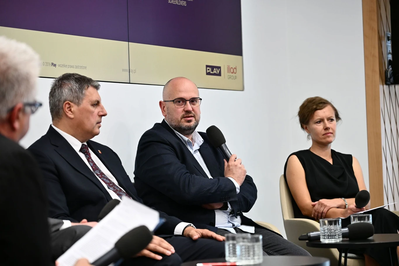 Cztery osoby siedzące przy stoliku podczas panelu dyskusyjnego, trzymające mikrofony; dwaj mężczyźni w garniturach oraz kobieta w czarnej sukience, skupieni na rozmowie.